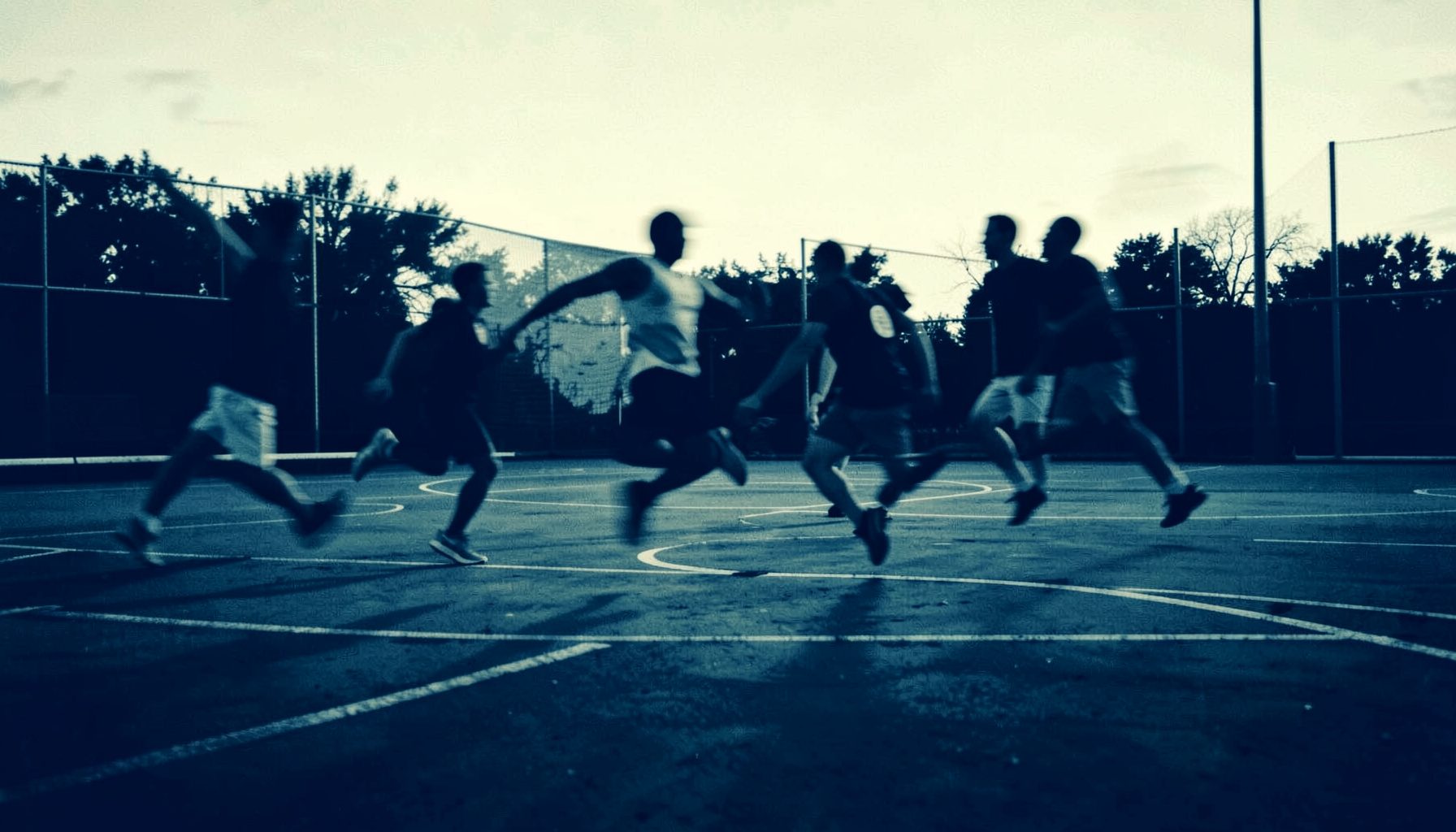 Stylized duotone photograph in deep navy and warm cream, showing an empty outdoor sports court at dusk with motion-blurred silhouettes of athletes leaping mid-play, conveying speed and casual street-sport energy.